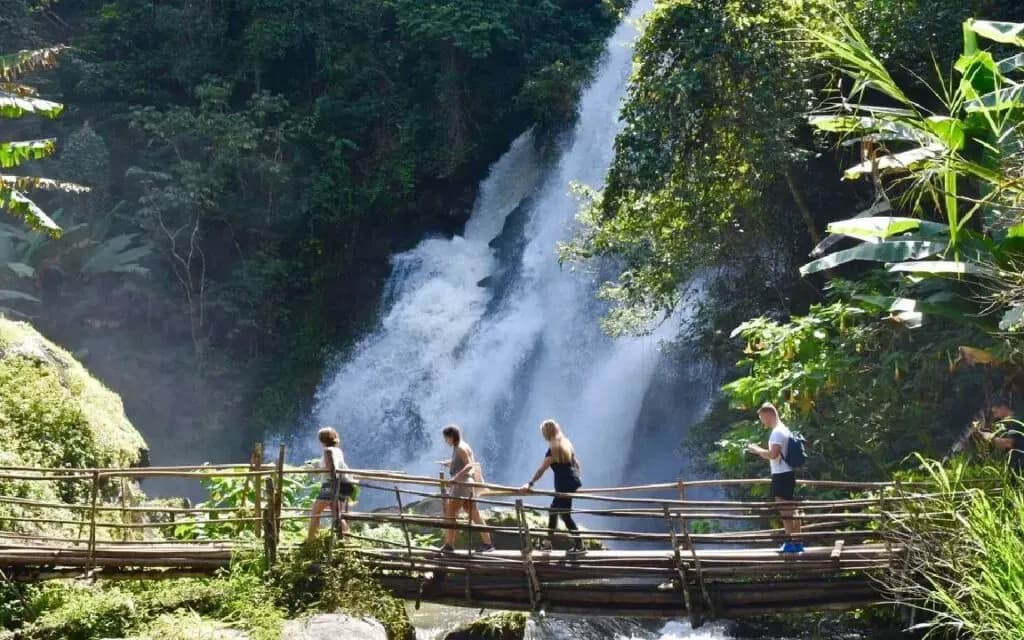 Tourists hiking at Pha Dok Siew Nature Trail, walking on a bamboo bridge with a waterfall cascading in the background
