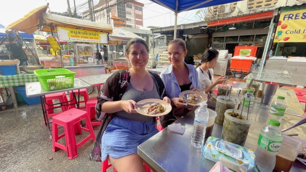 This image features two women enjoying local street food at a market in Chiang Mai. They are seated at a small, casual dining setup typical of Thai street food stalls, with various condiments on the table. The market environment, with other vendors and signage in the background, helps convey the bustling and authentic dining experience that tourists can expect in Chiang Mai. The purpose of this image is to highlight the affordable and authentic culinary experiences available to travelers, making it a great visual for promoting budget-friendly travel or local cuisine on a travel blog or website about Chiang Mai.
