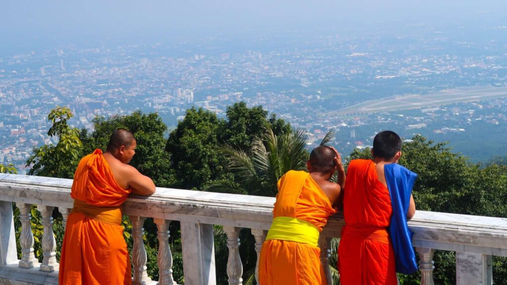 Three Buddhist monks admiring the vibrant Chiang Mai city skyline from the temple, a peaceful highlight of a Chiang Mai tour