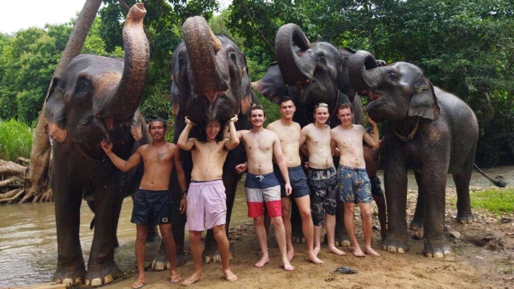Group of travelers enjoying a Chiang Mai tour at an elephant sanctuary, posing with elephants in a lush jungle near a river.