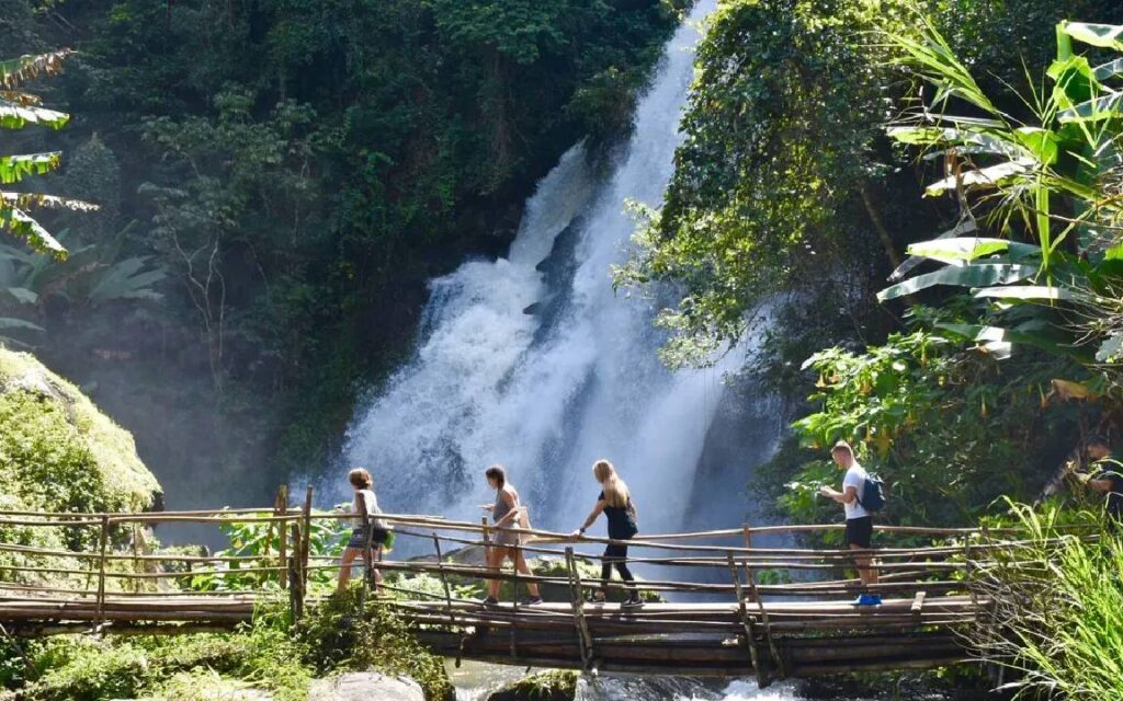 Tourists hiking at Pha Dok Siew Nature Trail, walking on a bamboo bridge with a waterfall cascading in the background